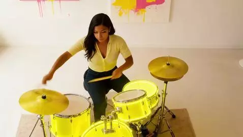 Drummer playing a neon yellow glowing drum kit with abstract paint splatters in the background.