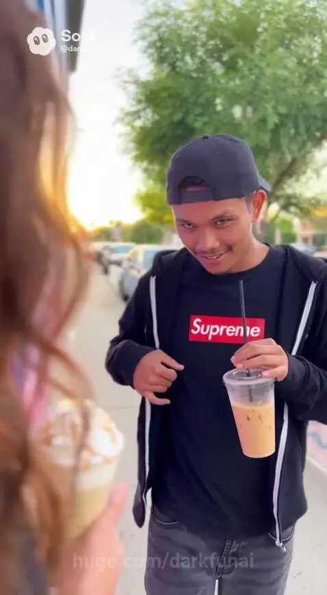 Man and woman walking outdoors, holding drinks, with the man about to demonstrate his accent for picking up girls.