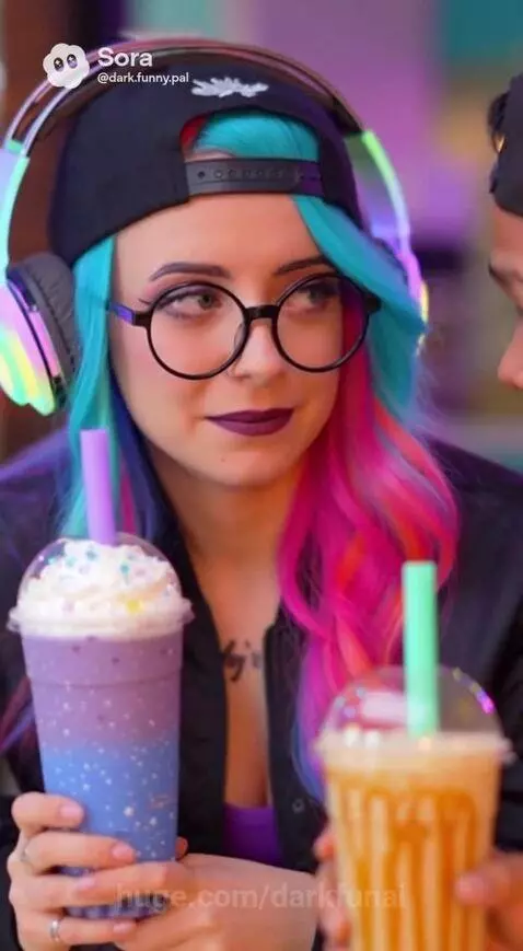 Young man and woman with blue/pink hair at a boba tea shop table, holding drinks.