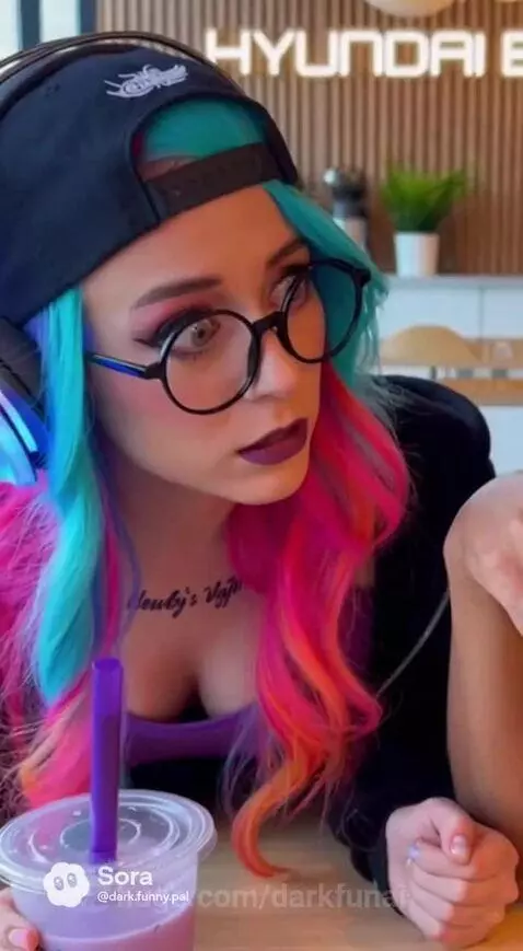 Man with blue and pink hair tells a joke while woman sips bubble tea; he spills it, and they laugh.