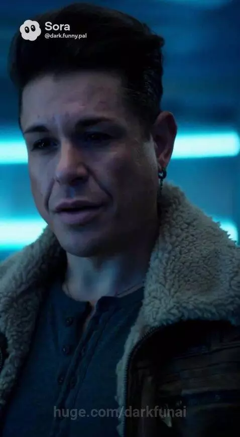 A blonde woman and a man in a fur-collared jacket look at a glowing blue cryo-pod containing a figure.