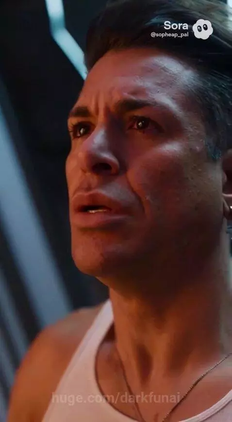 Man in white tank top distressed, questioning a green insect-like alien in a dimly lit futuristic corridor.