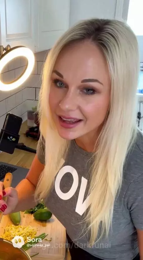 A blonde woman in a grey t-shirt and denim shorts smiles while stirring a pot in a kitchen, with a laptop showing a recipe.