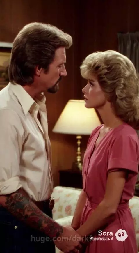 Couple holding hands and looking into each other's eyes in a living room, man with beard, woman with blonde curly hair.