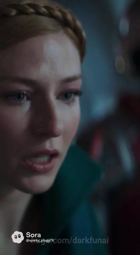 Woman with blonde braided hair crying, looking towards a glowing blue cylindrical chamber with a figure inside.