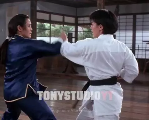 Two women in martial arts uniforms sparring intensely in a dojo, one in blue Kung Fu attire, the other in a white Karate gi.