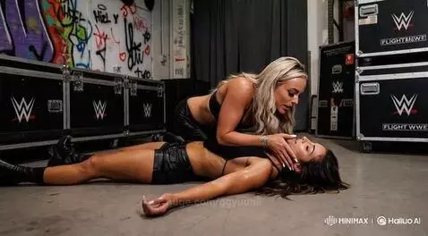 Blonde woman in black bra top kisses incapacitated brunette woman on the ground, WWE equipment visible.