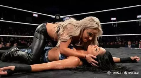 Woman with blonde hair in black outfit kneeling over woman with dark hair in blue top, leaning in for a kiss.