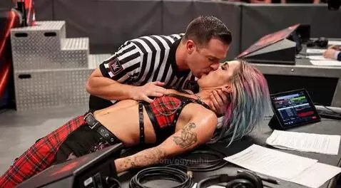 Referee in striped shirt kissing a woman with colorful hair and tattoos lying on a table.