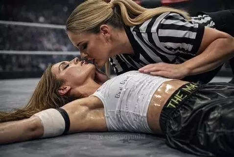 Referee leaning over and kissing a female wrestler lying on the mat in a wrestling ring.