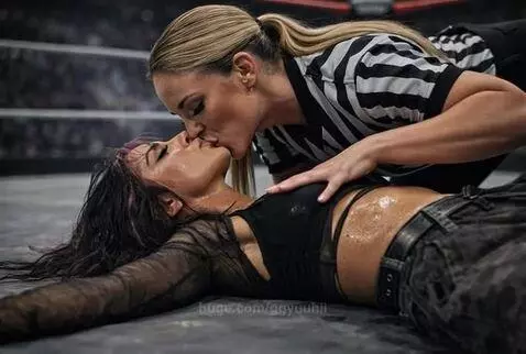 Wrestling referee with blonde ponytail kissing a woman with dark purple-streaked hair lying on the ground in a wrestling ring.