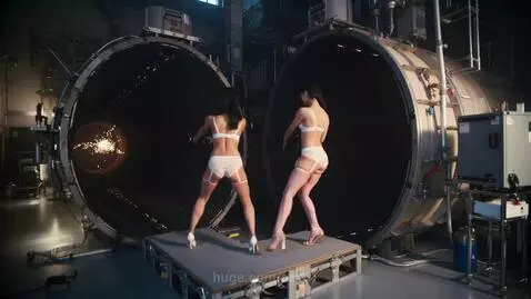 Two women in lingerie stand with their backs to the camera, aiming laser beams into dark cylindrical tunnels, creating sparks.