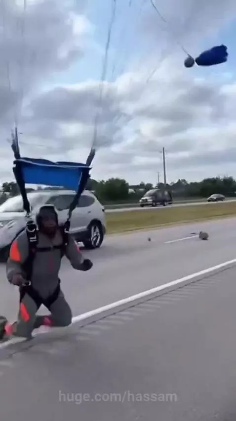 Skydiver in grey and orange jumpsuit landing on a highway median with parachute collapsing.