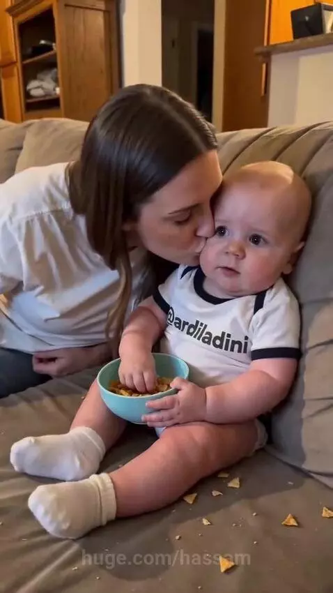 Baby eating snacks on couch, mom leans in for a kiss, baby looks away.