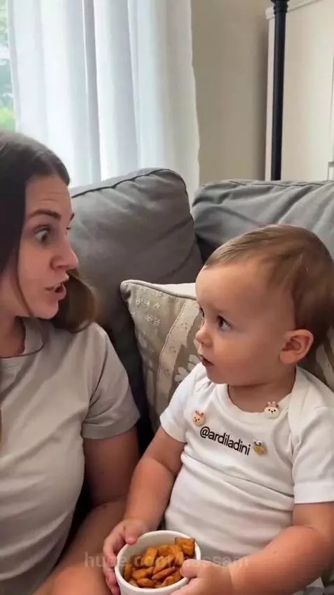 A baby sits on a couch holding a bowl of snacks and tells his mom he needs a wife to share them with.