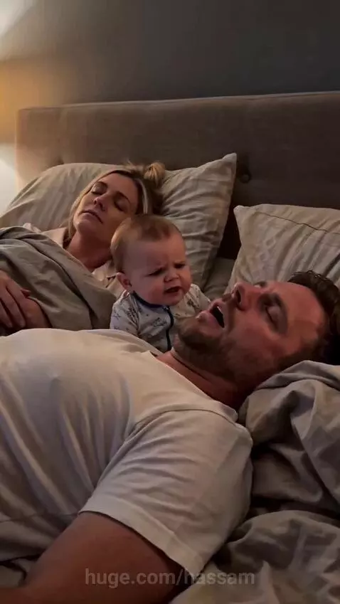 Close-up of an irritated baby looking at the camera, with a woman and a man sleeping in the background.
