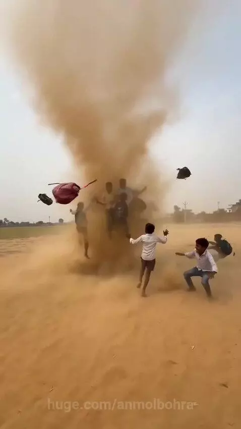 Young boys with backpacks are lifted and spun by a large dust devil in a sandy field.