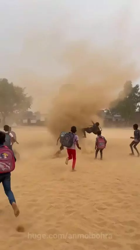 Young boys in school bags run through and are lifted by a large dust devil in a sandy field, appearing to have fun.