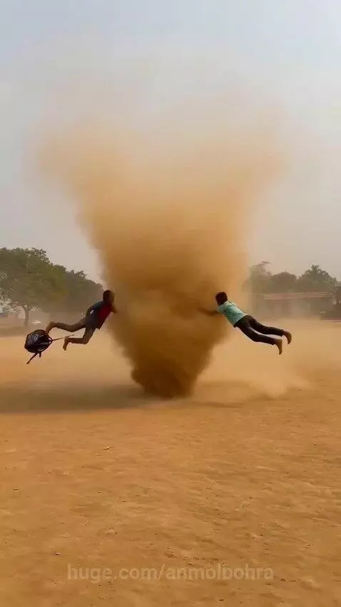 Young boys with backpacks being lifted by a dust devil in a dry, open field.
