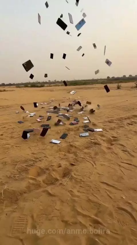 Numerous colorful smartphones falling and cracking on a vast, sandy landscape under a hazy sky.