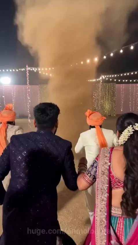 Guests in traditional Indian attire joyfully interact with a dust devil at an outdoor night wedding event.