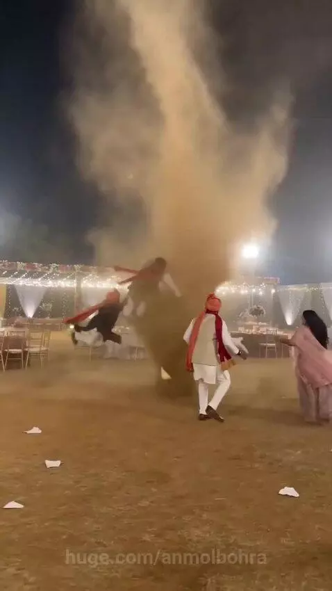 Men in traditional Indian attire are lifted by a large dust devil at a nighttime wedding reception.