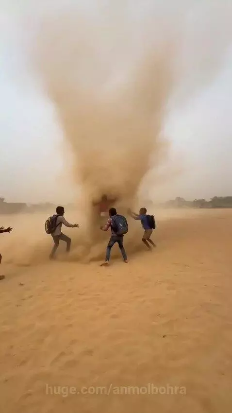 Several young boys with backpacks are lifted into the air by a large swirling dust devil in a sandy field.