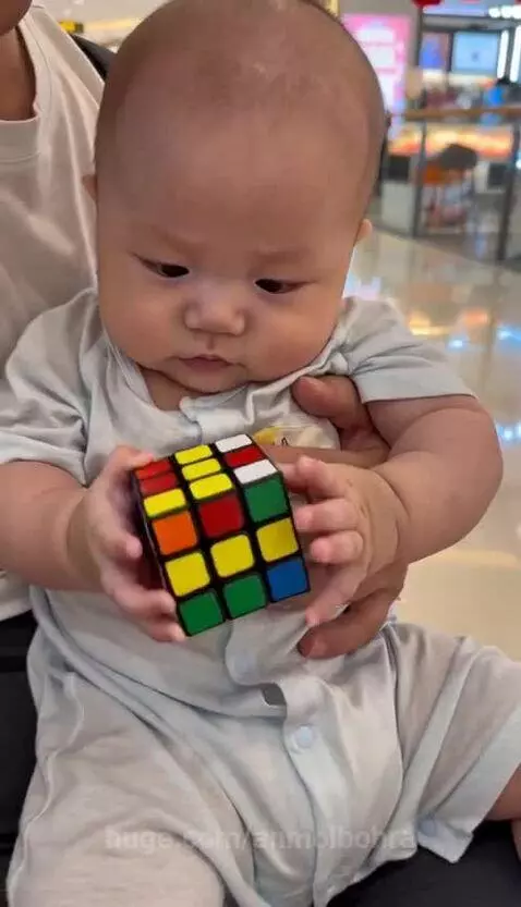 A baby in a light onesie focuses on a Rubik's cube, held by an adult hand, then laughs joyfully while playing with it.