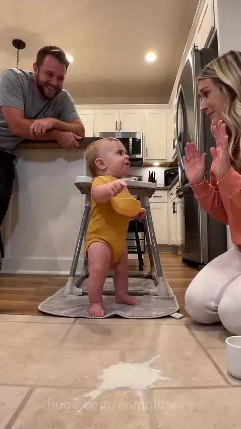 Baby in a yellow onesie standing in a high chair, looking at a milk spill on the floor while parents clean.
