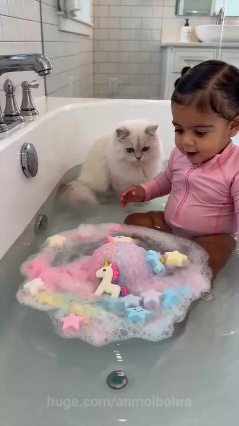 Baby laughing with excitement as a colorful unicorn bath bomb fizzes in the tub, with a cat nearby.