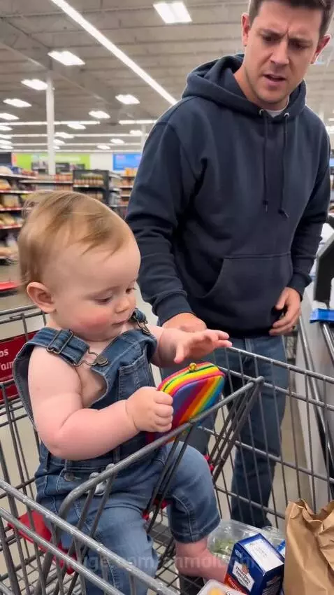 Baby in a shopping cart holds a toy bill and wallet, handing it to a smiling cashier while dad laughs.