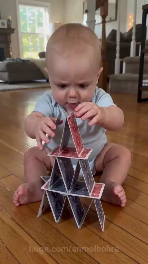 Baby sitting on floor, looking at collapsed card tower after sneezing.