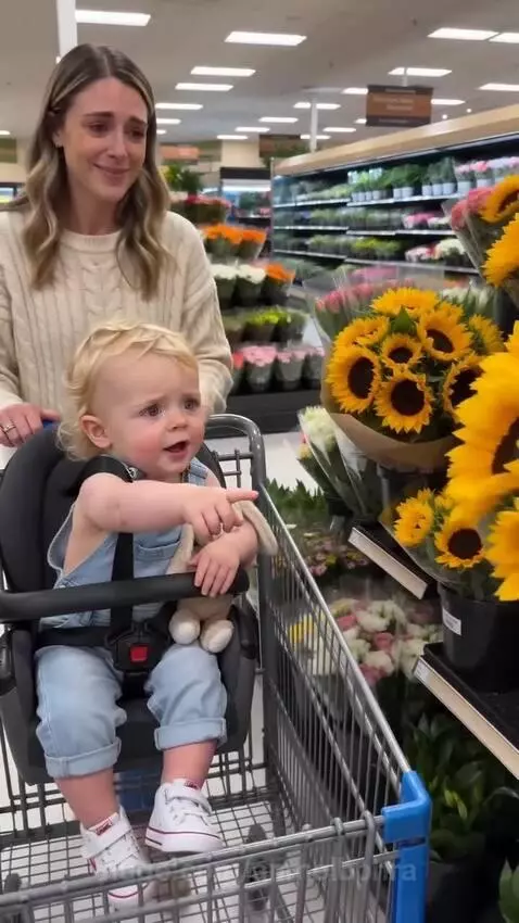 Toddler proudly holds a sunflower and smiles at his mother in a grocery store.