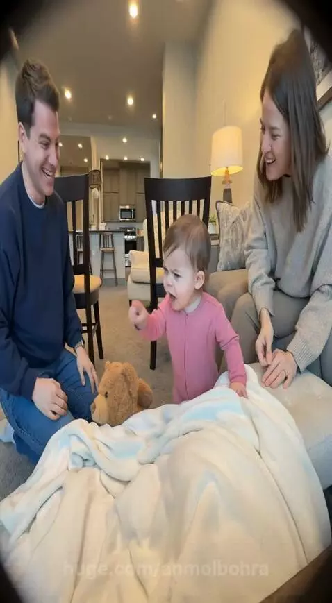 Baby in pink onesie directing parents as they build a blanket fort, with a dog nearby.