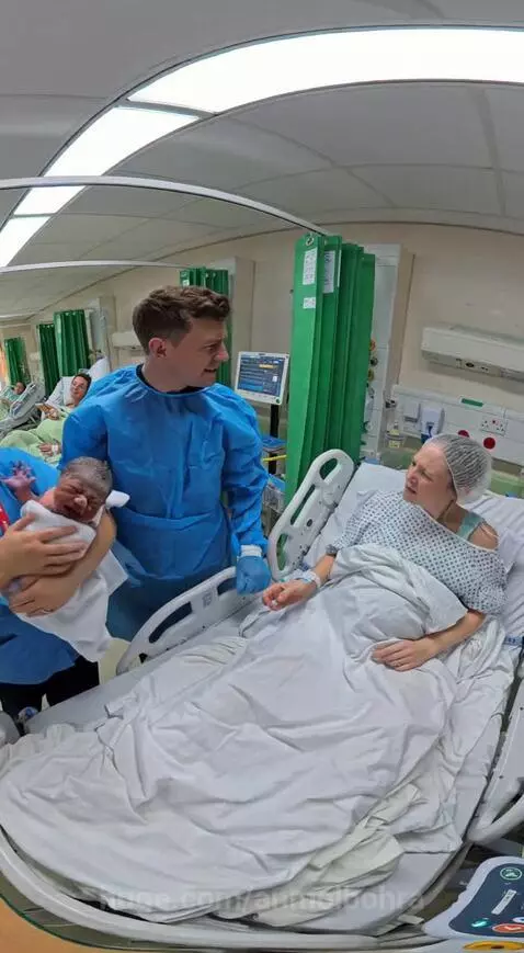 Dad looking confused at a newborn baby, with a nurse and mother present in a hospital room.
