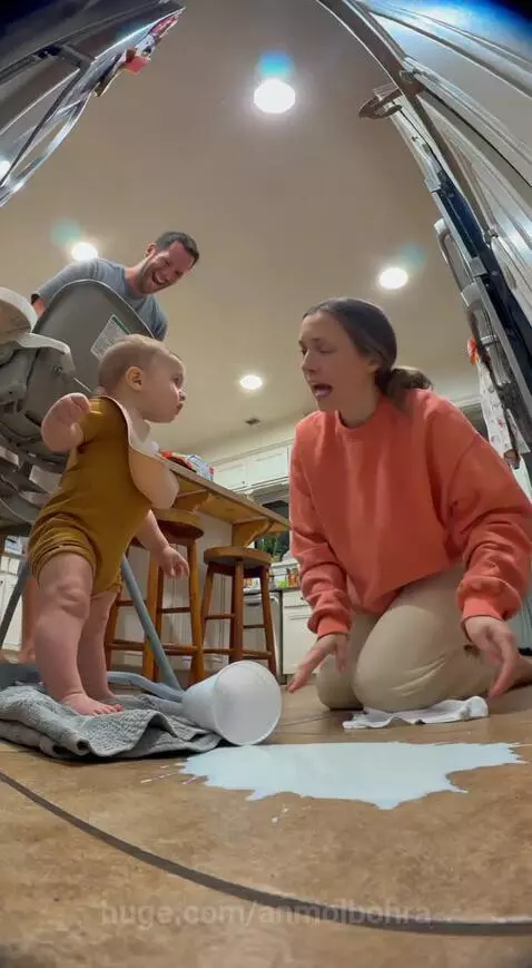 Baby in high chair points at spilled milk on kitchen floor while mom cleans and dad laughs.