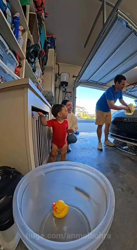 Baby in red onesie interacting with parents over a rubber duck during a car wash in a garage.