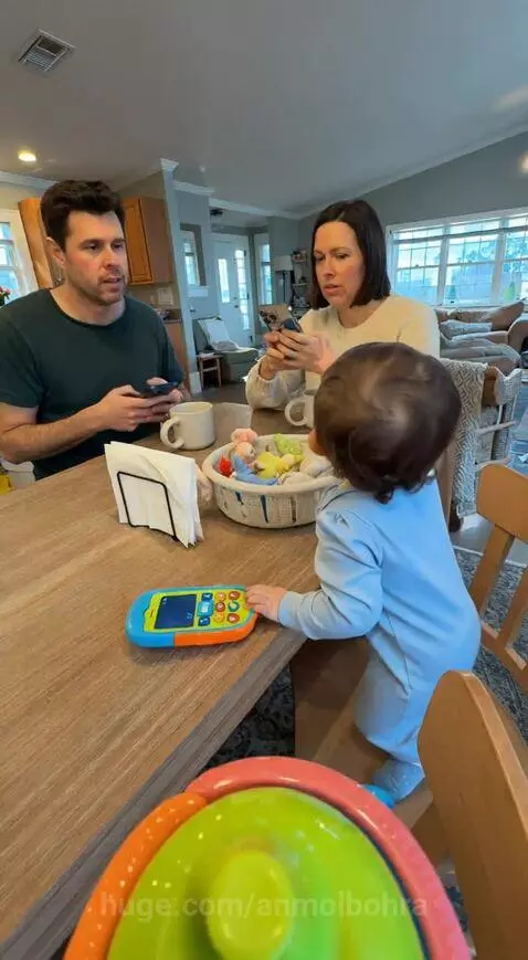 Baby in a blue onesie at a dining table, looking at the camera while parents put down their phones.