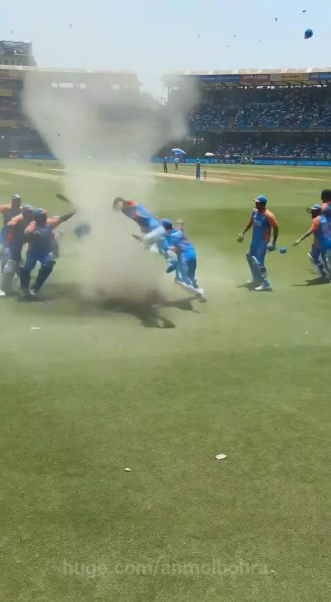 Cricket players in blue and orange uniforms tossed around by a dust devil on a field, losing hats.