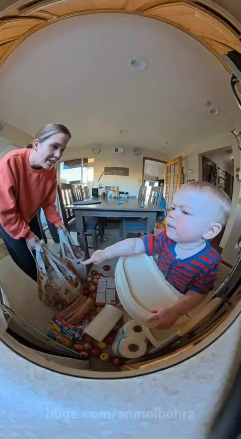 Baby in high chair points at spilled groceries on the floor while parents clean up.