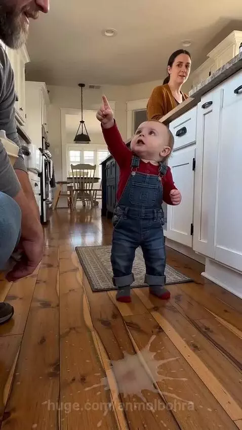 Baby in overalls and red shirt points at a spill on a wooden floor, then crosses arms with a disappointed look.