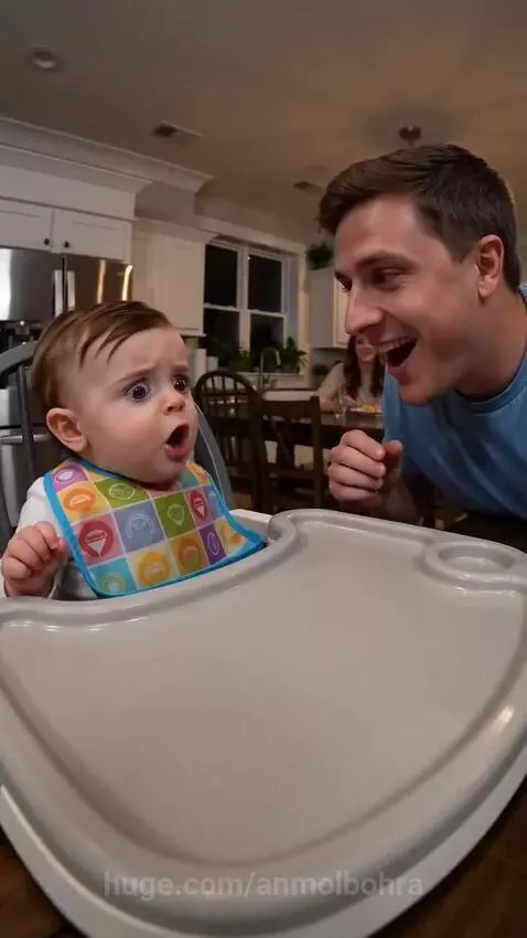 Baby in high chair with colorful bib, wide-eyed, pointing at dad who is pretending to eat their french fry.