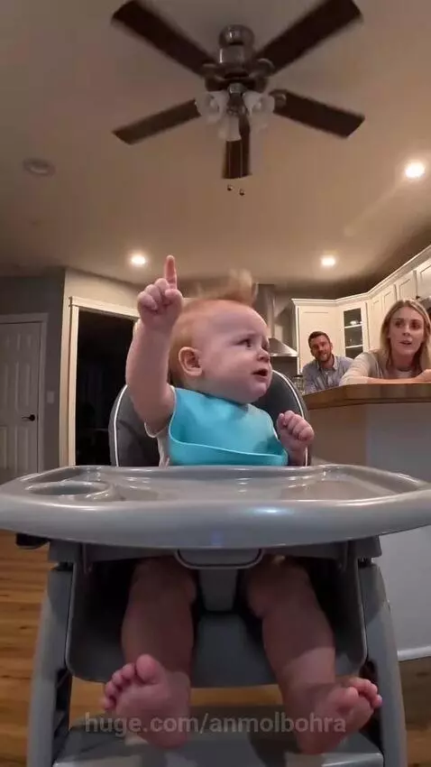 Baby in a high chair wearing a bib, pointing up and saying "no, no, no" and "I know".