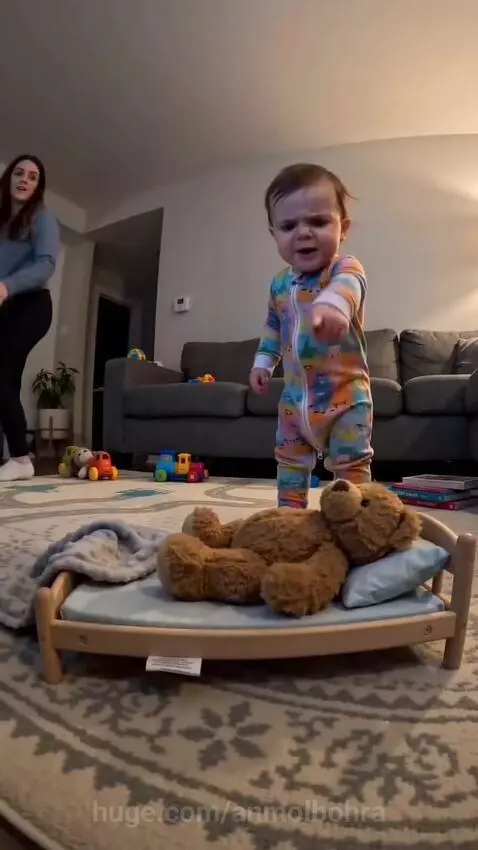 Baby in a colorful onesie stands by a bed and points, instructing a woman to cover a teddy bear.