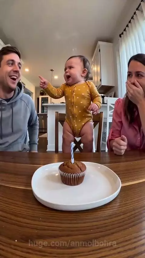 Baby in polka-dot onesie on a chair, facing a birthday cupcake with a lit candle, with parents watching.