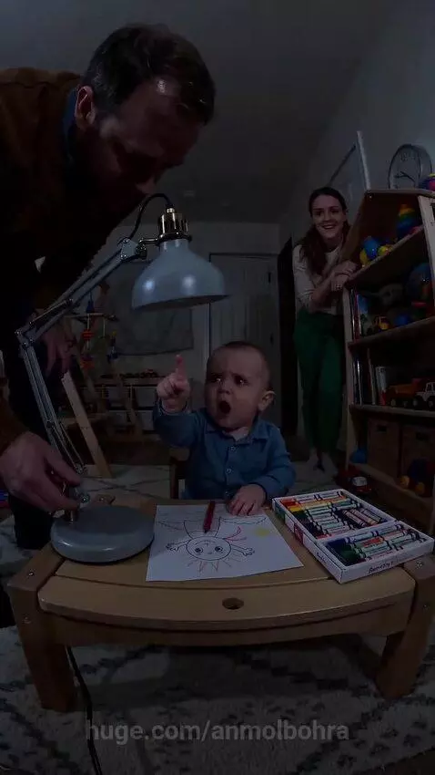 Baby coloring at a table, looking annoyed as a lamp is adjusted.