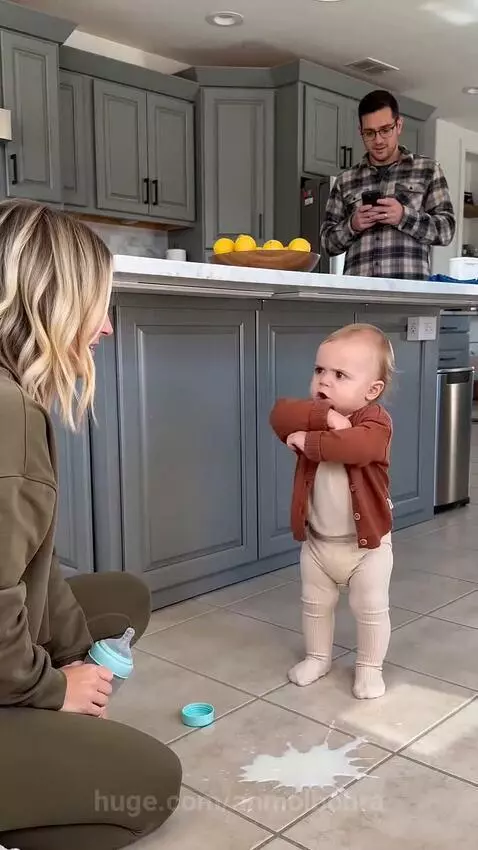 Baby in a white outfit and rust cardigan points at spilled milk on kitchen floor, scolding mom who is kneeling.