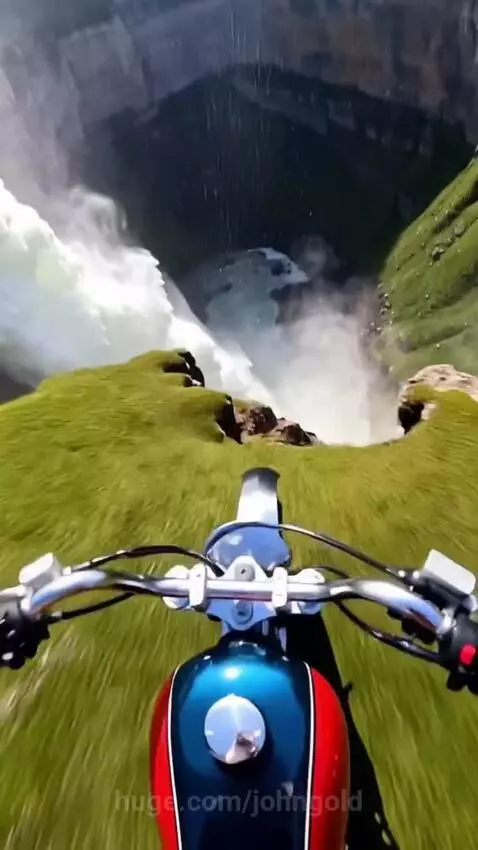 First-person view of a motorcycle driving off a cliff towards a giant waterfall in a lush canyon.