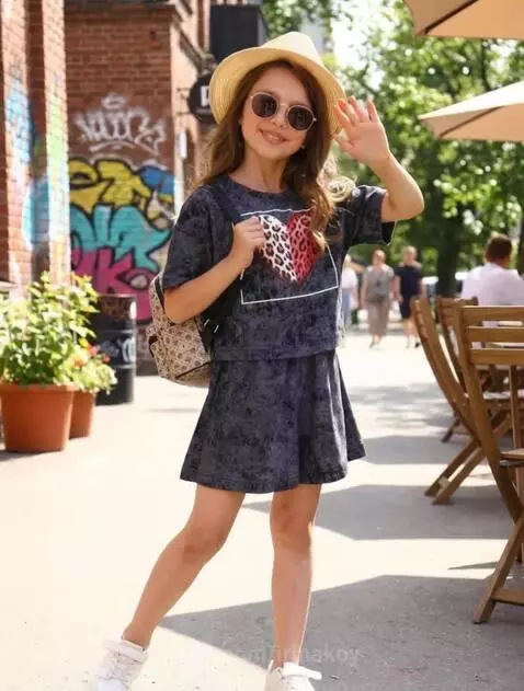 Young girl in a straw hat and sunglasses smiles and waves outdoors with a graffiti wall and cafe in the background.