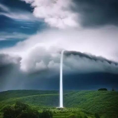 Massive luminous water column descending from a dark storm cloud to a green landscape.